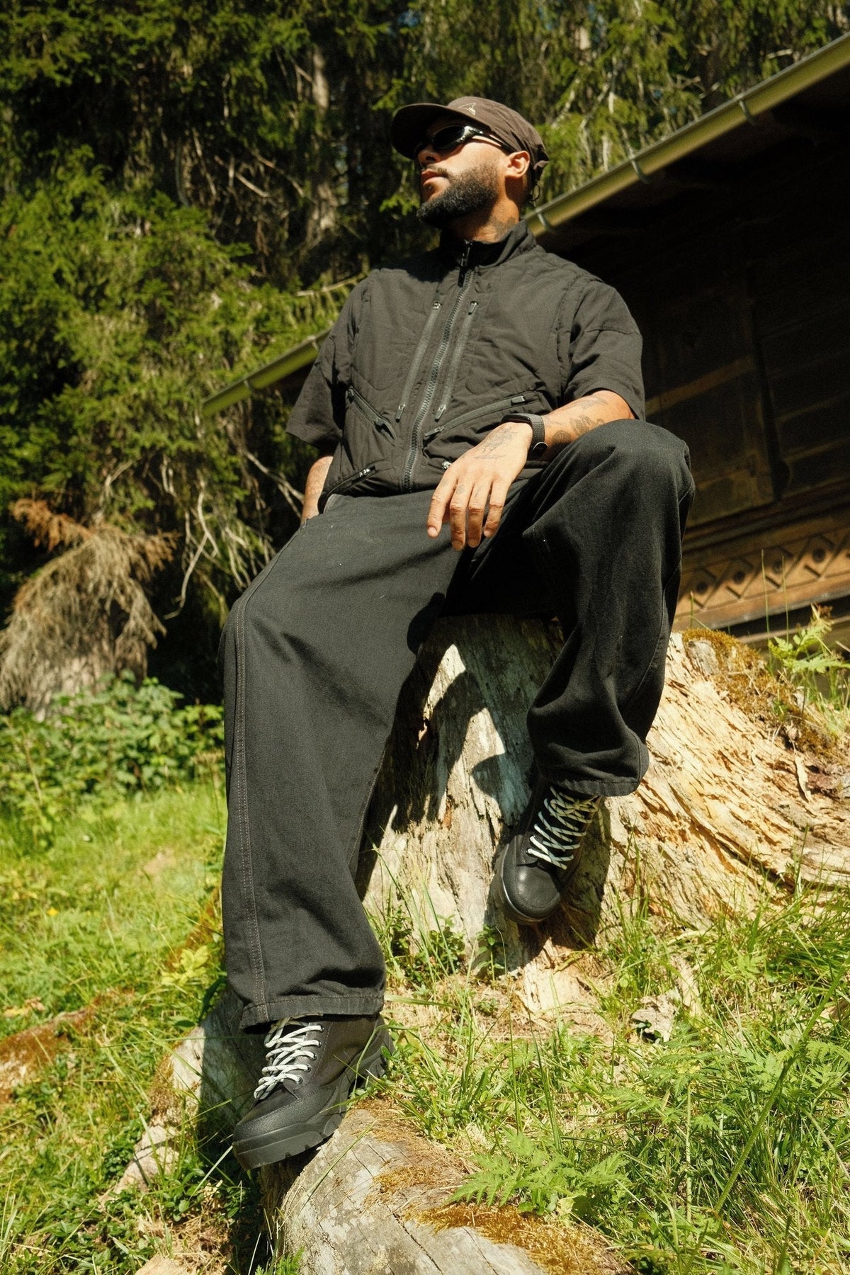 Man in black seated on a log outside an alpine chalet, wearing the Paper Straight Alpine Hiker in Black